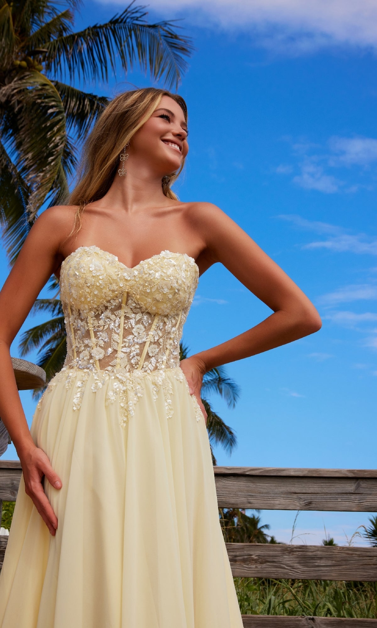Wearing the Nox Anabel C1617 Strapless Sweetheart Prom Dress, a woman stands outdoors by a wooden railing with palm trees and blue sky, her light yellow gown’s floral lace bodice perfectly suited to the sunny, elegant setting.